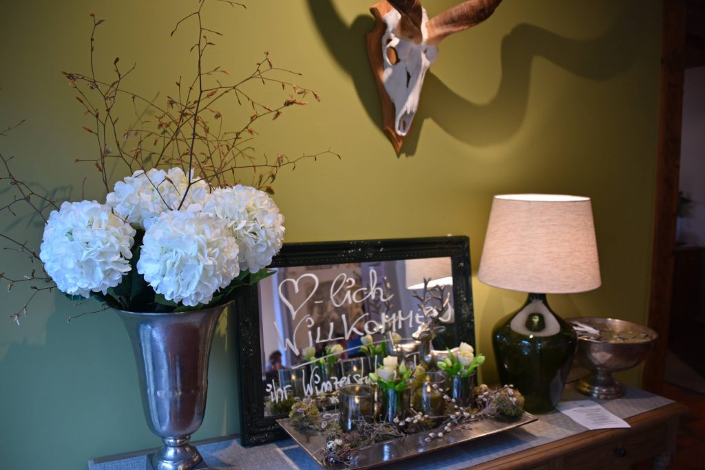 Das Bild zeigt ein dekoratives Arrangement auf einer Holzkommode vor olivgrüner Wand. Links steht ein großer silberner Pokal mit weißen Hortensien und Zweigen, daneben ein Spiegel mit der handgeschriebenen Begrüßung „Herzlich Willkommen“. Davor ein Tablett mit Glasvasen, Frühlingsblumen und Naturmaterialien. Rechts eine grüne Tischlampe mit beigem Schirm sowie eine silberne Schale. An der Wand hängt ein Geweih mit Schädel auf Holzplatte.